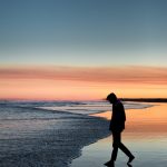silhouette of man walking on seashore under orange sky